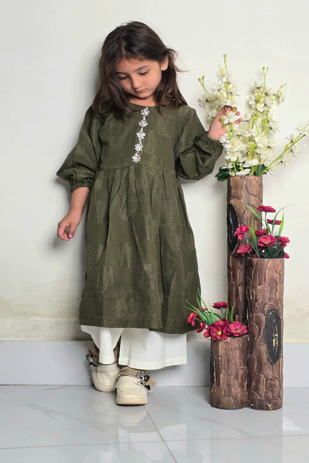 Young girl in an olive green dress with white floral details standing next to a wooden vase with flowers on a white floor.