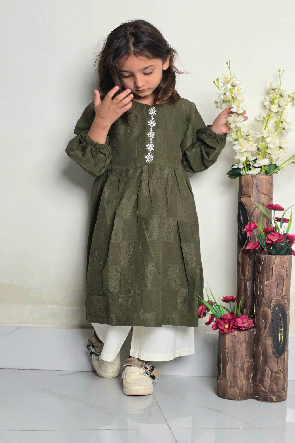 Young girl in a green dress standing next to a wooden vase with flowers on a white floor.