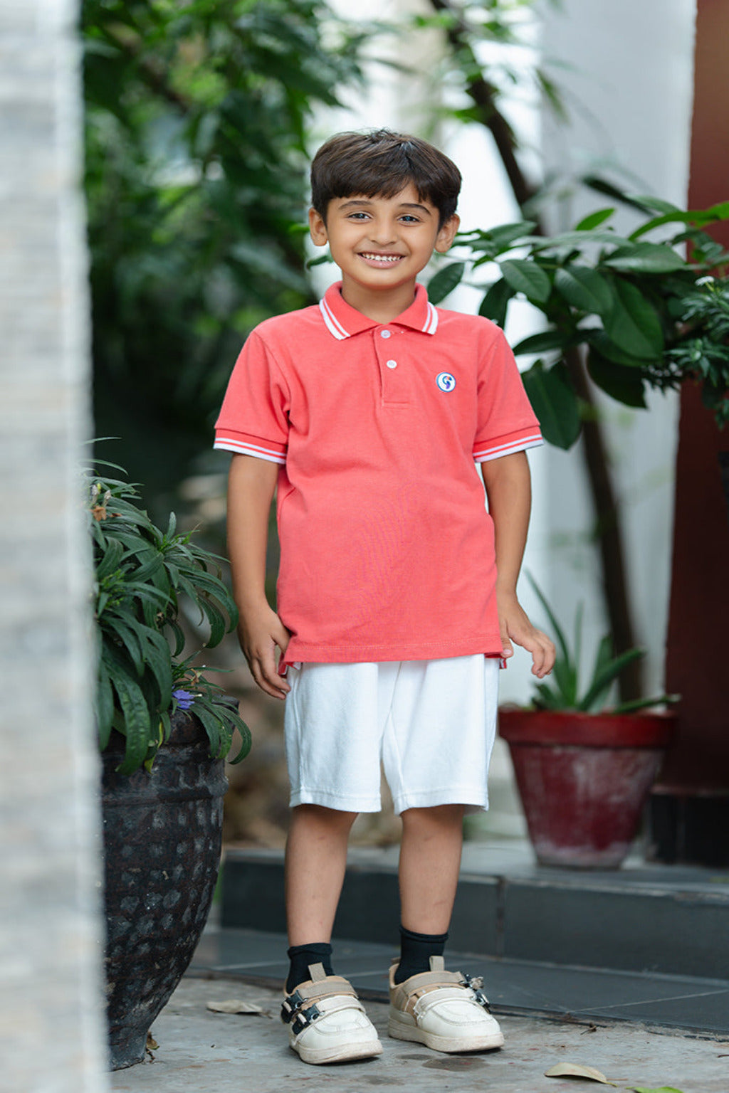 Young boy wearing a red polo shirt and white shorts standing outdoors with plants in the background