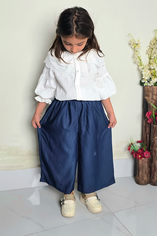 Young girl wearing a white blouse and blue pants standing indoors with a neutral background