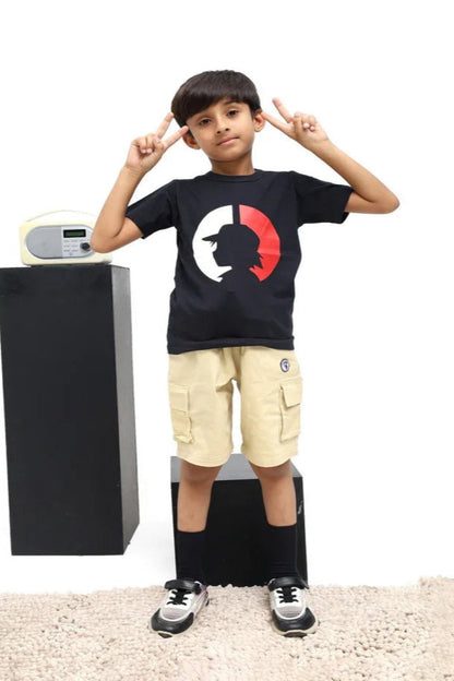 Child wearing a black t-shirt with a red and white design, beige cargo shorts, and black sneakers, standing next to a black speaker on a light-colored floor.