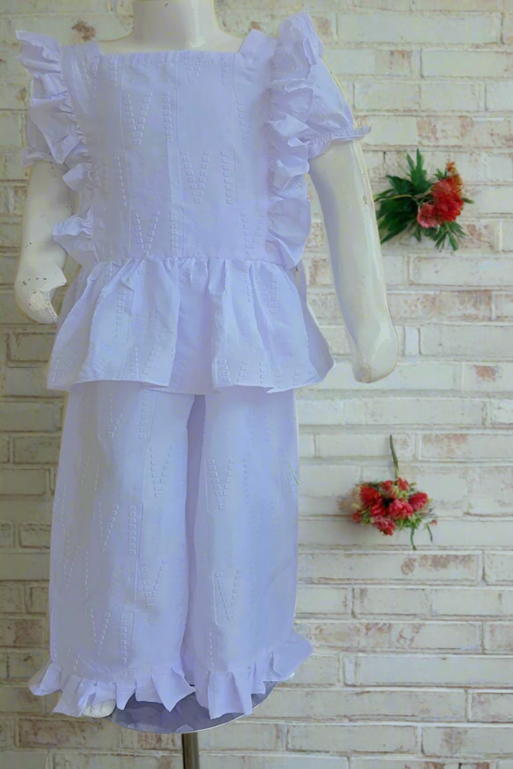 White dress with ruffled details on a mannequin against a white brick wall with floral decorations.