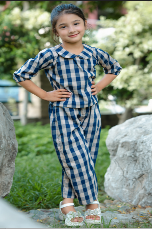 Young girl in a blue and white checkered outfit standing outdoors with greenery in the background