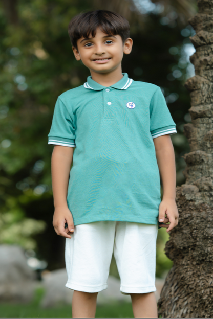 Child wearing a teal polo shirt and white shorts standing outdoors with greenery in the background