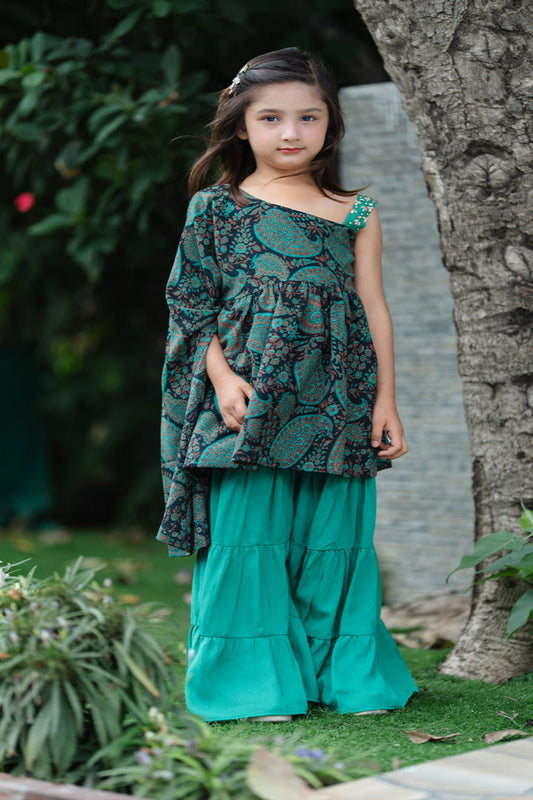 Young girl in a green and black patterned outfit standing outdoors near a tree