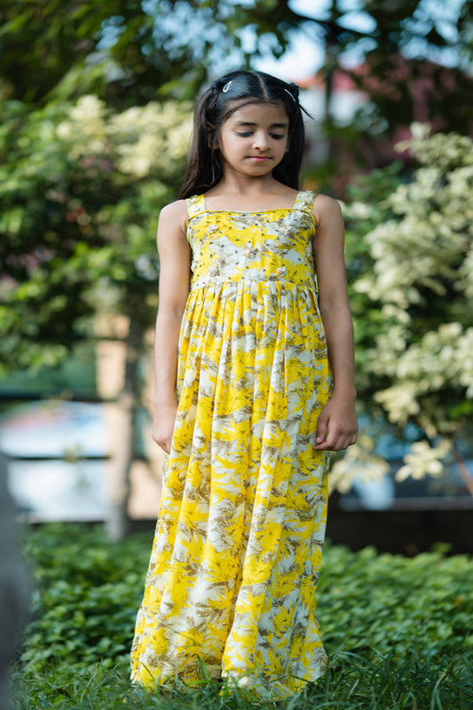 Young girl in a yellow floral dress standing outdoors with greenery in the background